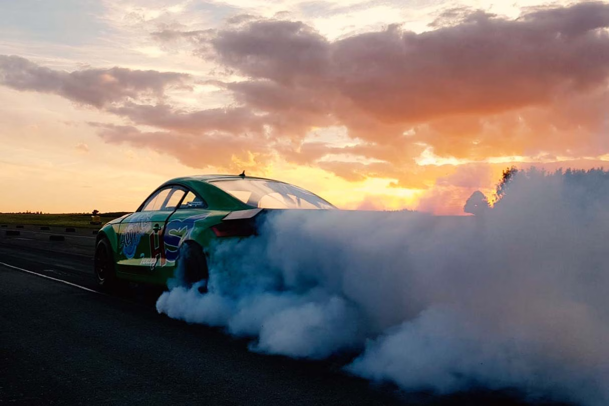 Grünes HST-Tuning-Auto beim Burnout auf der Rennstrecke, dichter Rauch steigt auf, Sonnenuntergang im Hintergrund.