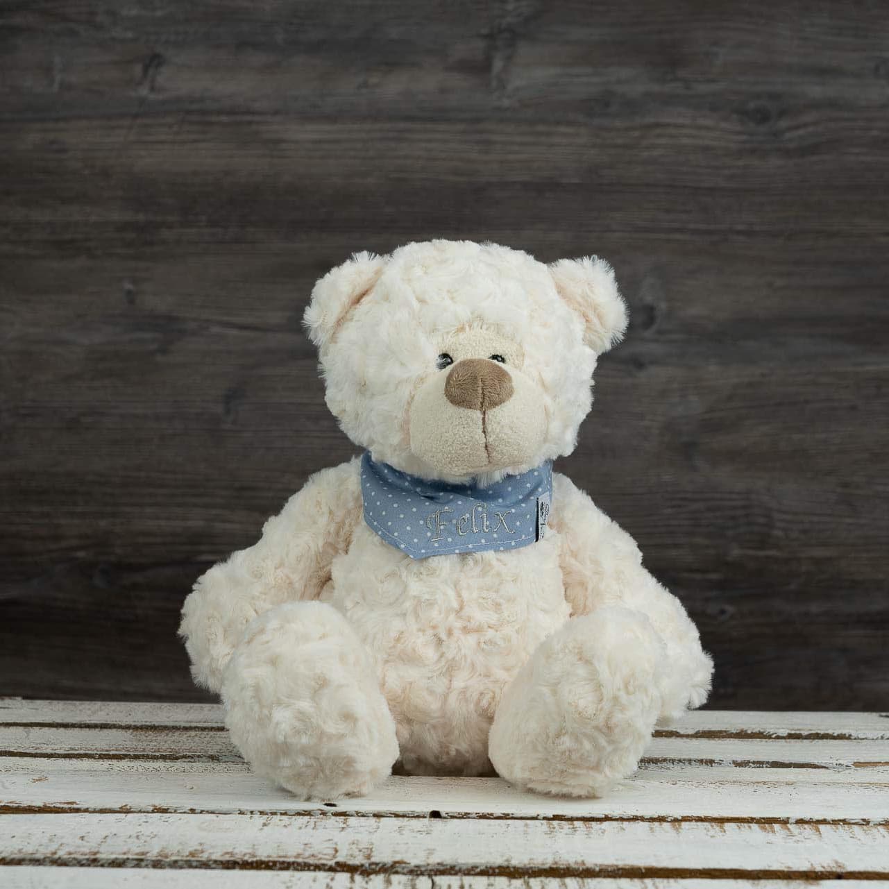 Weißer Teddybär mit blauer Halstuch-Schleife und „Felix“-Stickerei auf rustikalem Holztisch.