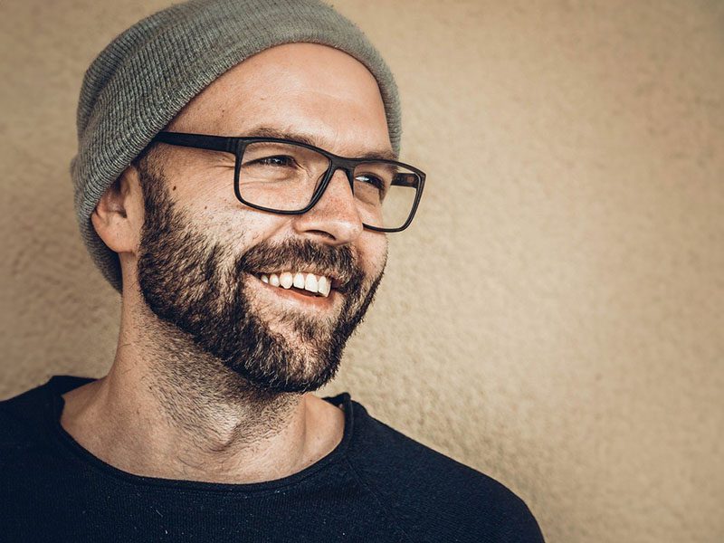 Porträt eines herzlich lachenden Mannes mit Vollbart, schwarzer Brille und grauer Mütze, der zur Seite blickt