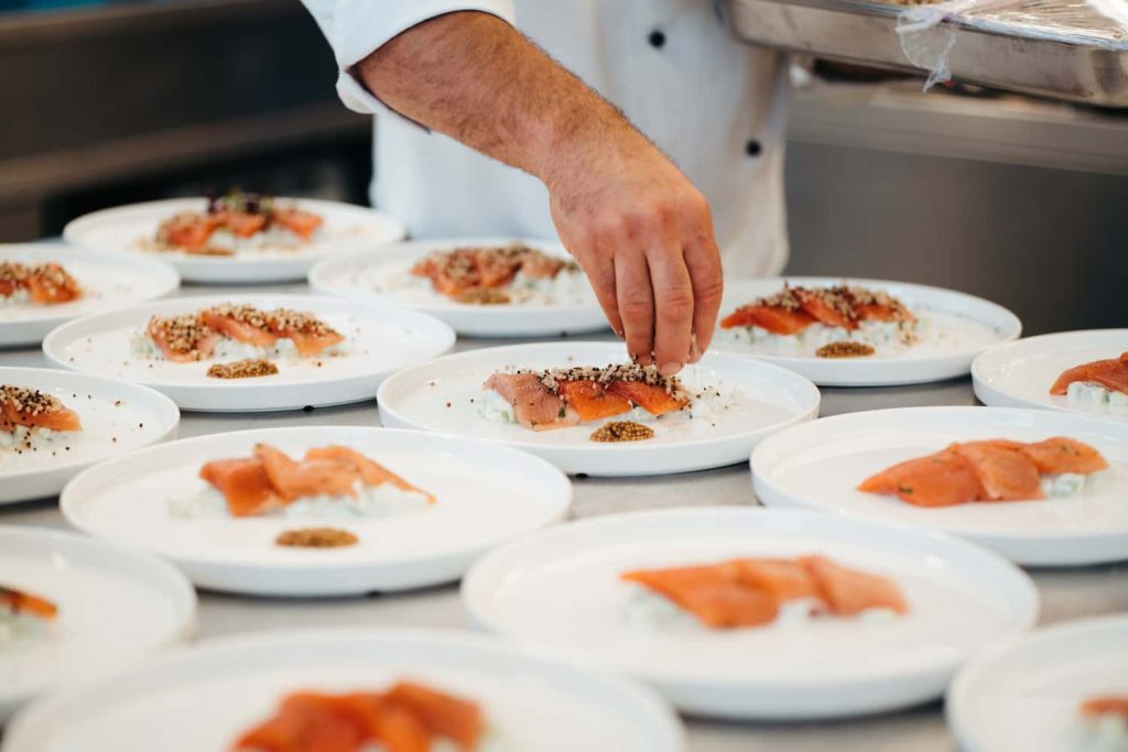 Adventure Catering Performance Marketing 3 Vorspeise von Adventure Catering: ein Koch richtet Lachshäppchen auf weißen Tellern mit feinen Toppings und Beilagen an.