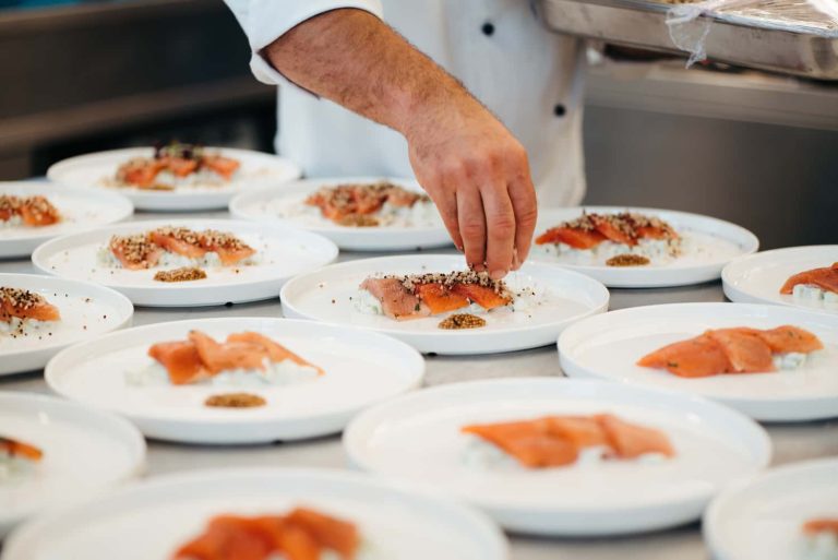 Vorspeise von Adventure Catering: ein Koch richtet Lachshäppchen auf weißen Tellern mit feinen Toppings und Beilagen an.