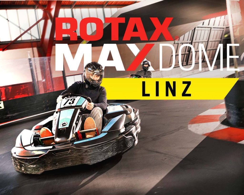 Racing Fieber im Rotax MAX Dome 1 Fahrer im E-Kart auf Indoor-Strecke, darüber Schriftzug „ROTAX MAX DOME LINZ“ in rot, weiß und gelb vor Hallenkulisse.
