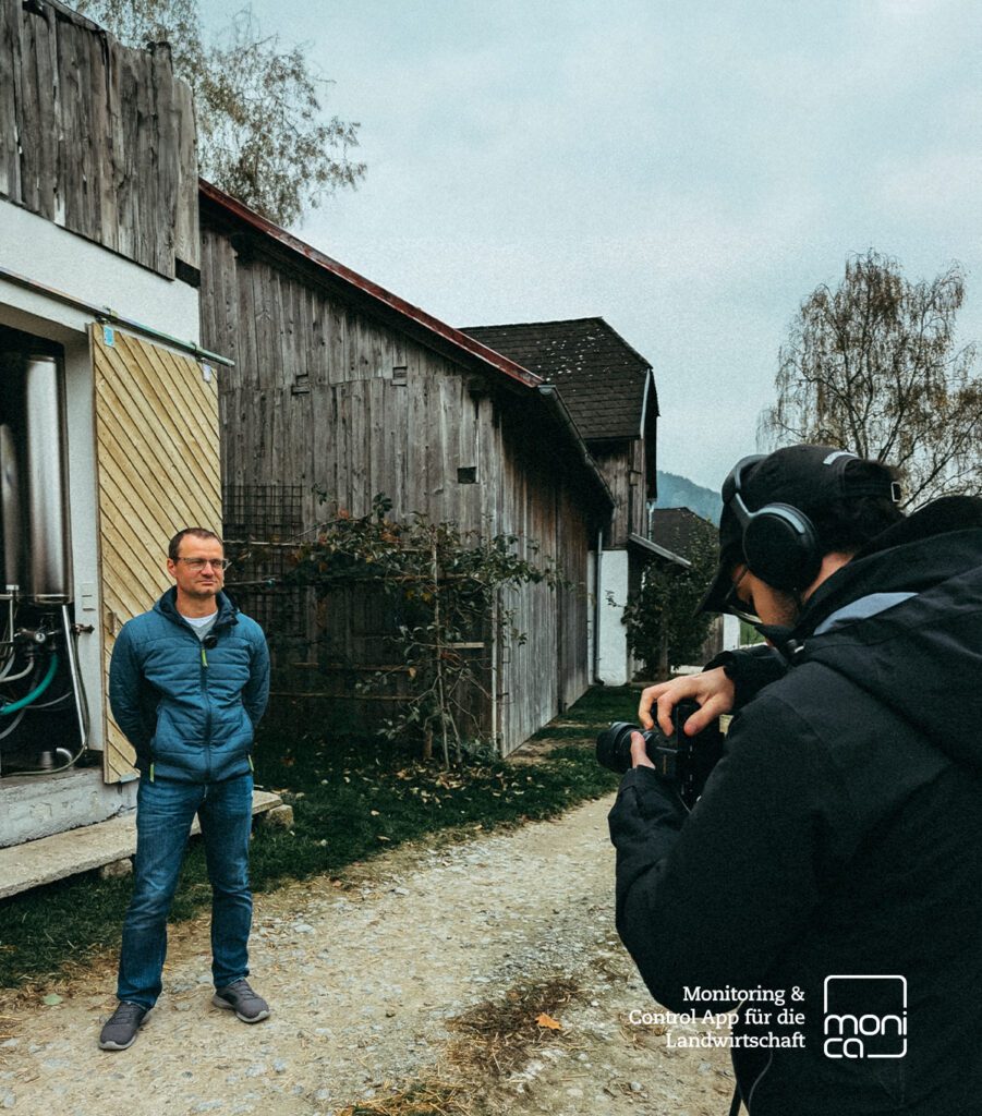 Landwirt steht vor Stallgebäude, wird von Person mit Kamera und Kopfhörer gefilmt, Logo der Landwirtschafts-App „monica“ unten rechts
