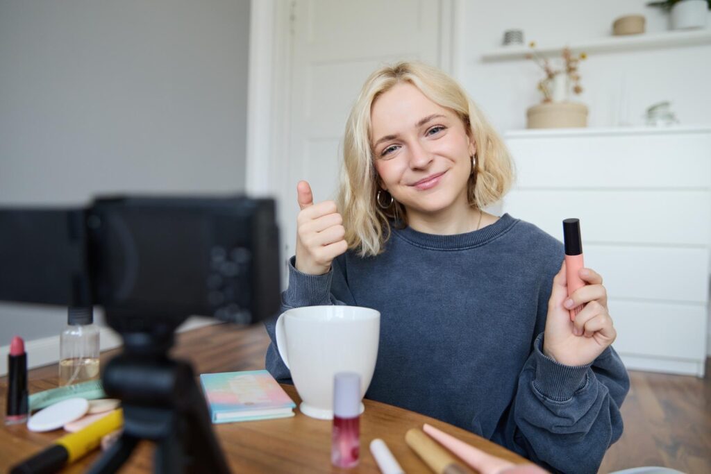 Junge Frau (Influencerin), welche an einem Tisch vor einer Kamera sitzt, lächelt und den Daumen nach oben hält, während sie in der anderen Hand ein Makeup-Produkt in die Kamera hält. 